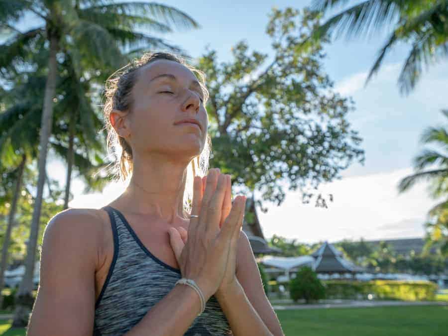 Eine Frau in Namaste-Pose vor tropischer Kulisse – inszeniert als Symbol für Achtsamkeit und Spiritualität