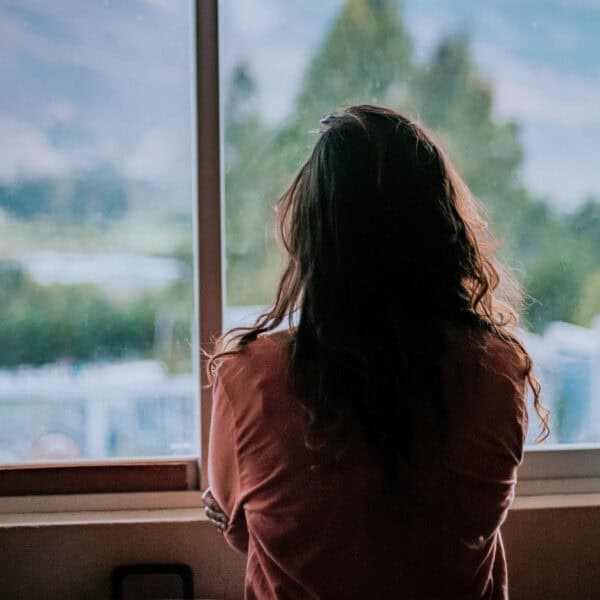 Frau steht mit dem Rücken zur Kamera vor einem Fenster und blickt nach draußen – Symbolbild für Zweifel und stille Erwartungen rund um Manifestation.