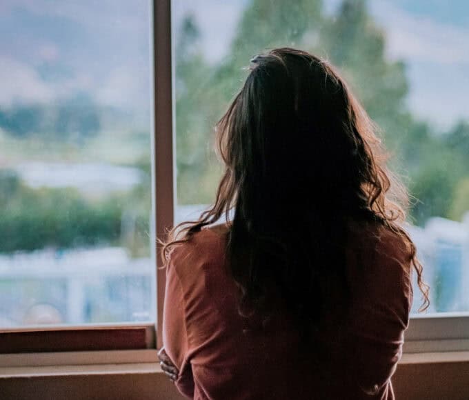 Frau steht mit dem Rücken zur Kamera vor einem Fenster und blickt nach draußen – Symbolbild für Zweifel und stille Erwartungen rund um Manifestation.