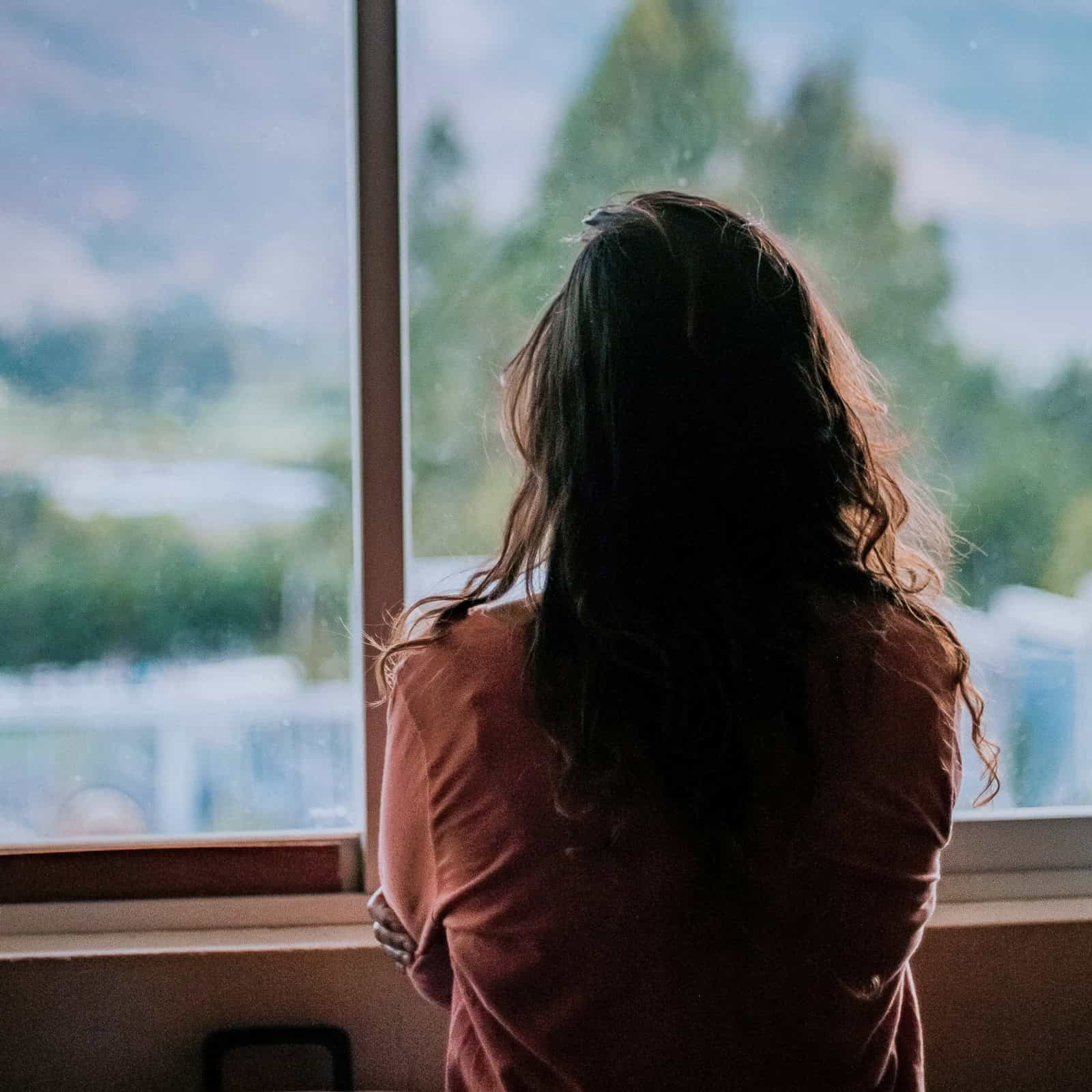 Frau steht mit dem Rücken zur Kamera vor einem Fenster und blickt nach draußen – Symbolbild für Zweifel und stille Erwartungen rund um Manifestation.