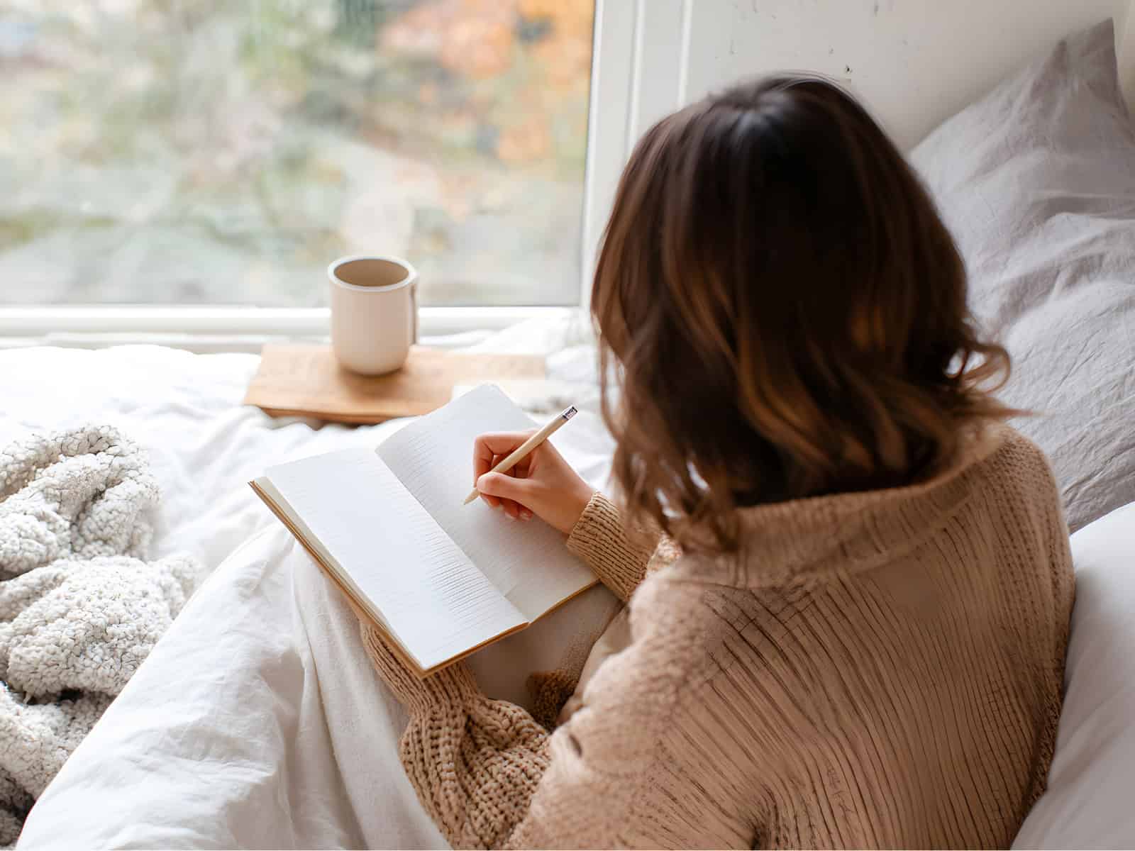 Frau sitzt auf einem Bett und schreibt in ein Notizbuch, neben ihr steht eine Tasse Kaffee am Fenster.