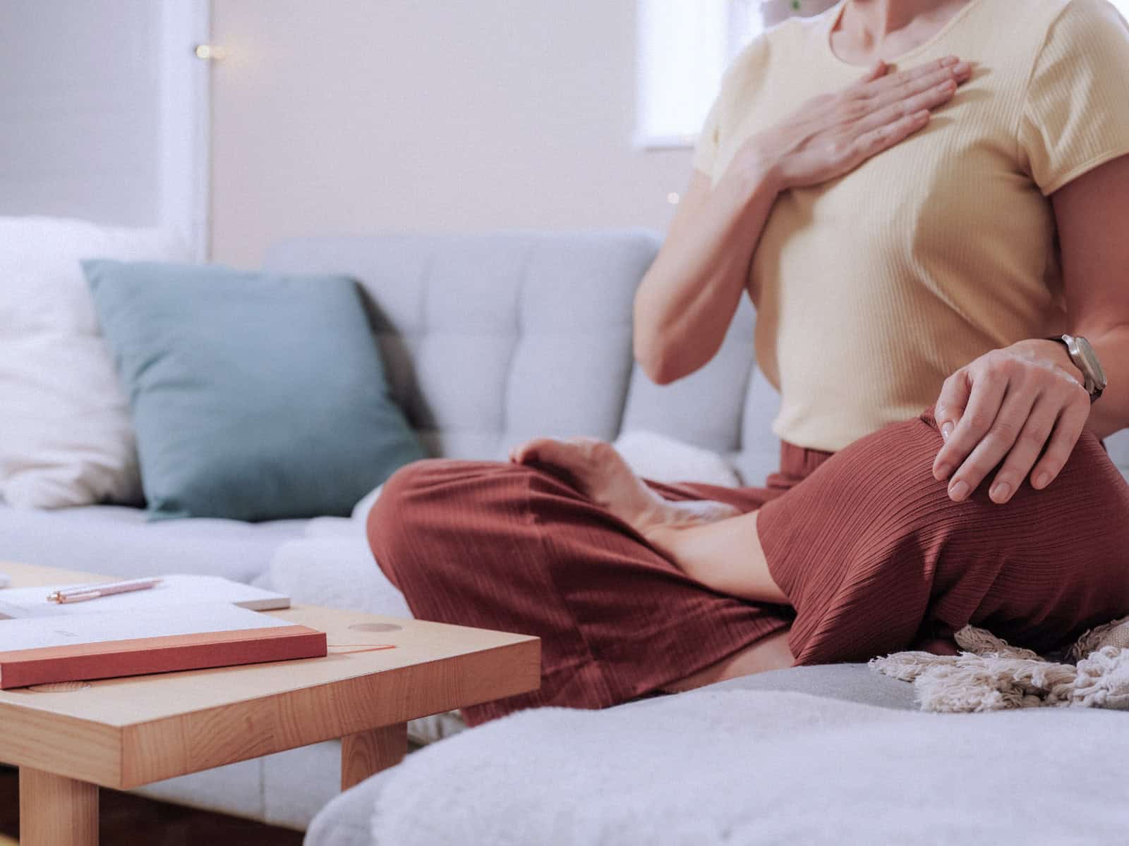 Manifestation - Frau sitzt im Schneidersitz auf einem Sofa und schreibt in ein Notizbuch, vor ihr liegt ein Journal auf dem Tisch.