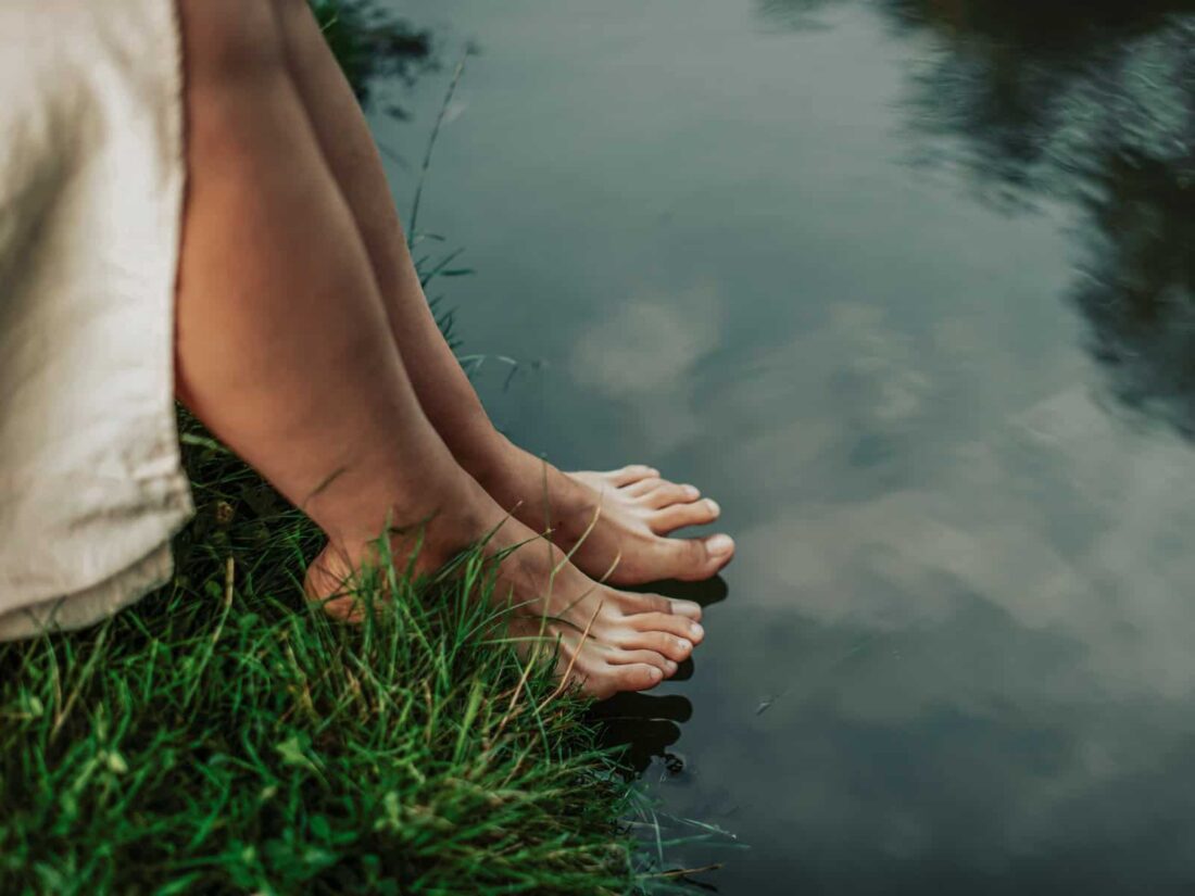Echte Spiritualität: Barfuß stehende Füße am Ufer eines ruhigen Gewässers, Gras berührt die Haut, das Wasser spiegelt den Himmel.