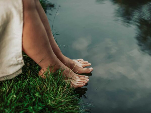 Echte Spiritualität: Barfuß stehende Füße am Ufer eines ruhigen Gewässers, Gras berührt die Haut, das Wasser spiegelt den Himmel.