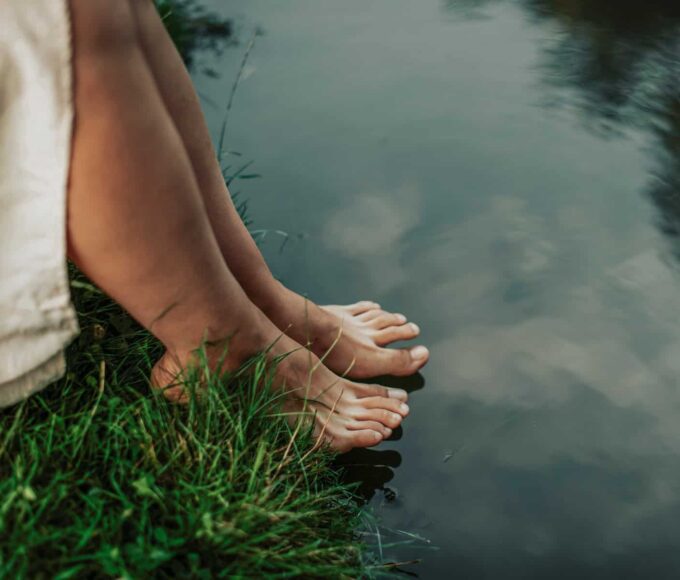 Klassische Spiritualität: Barfuß stehende Füße am Ufer eines ruhigen Gewässers, Gras berührt die Haut, das Wasser spiegelt den Himmel.