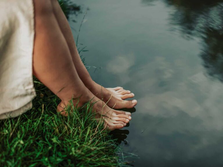 Klassische Spiritualität: Barfuß stehende Füße am Ufer eines ruhigen Gewässers, Gras berührt die Haut, das Wasser spiegelt den Himmel.