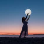 Frau hält leuchtenden Mond am Strand bei Sonnenuntergang – symbolisiert die emotionale Anziehung der Astrologie