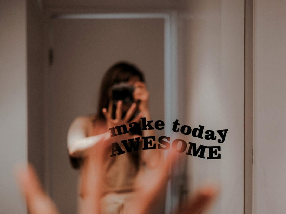 Spiegel mit der Aufschrift „make today awesome“, darin unscharf eine Person mit Smartphone in einem Wohnraum gespiegelt.