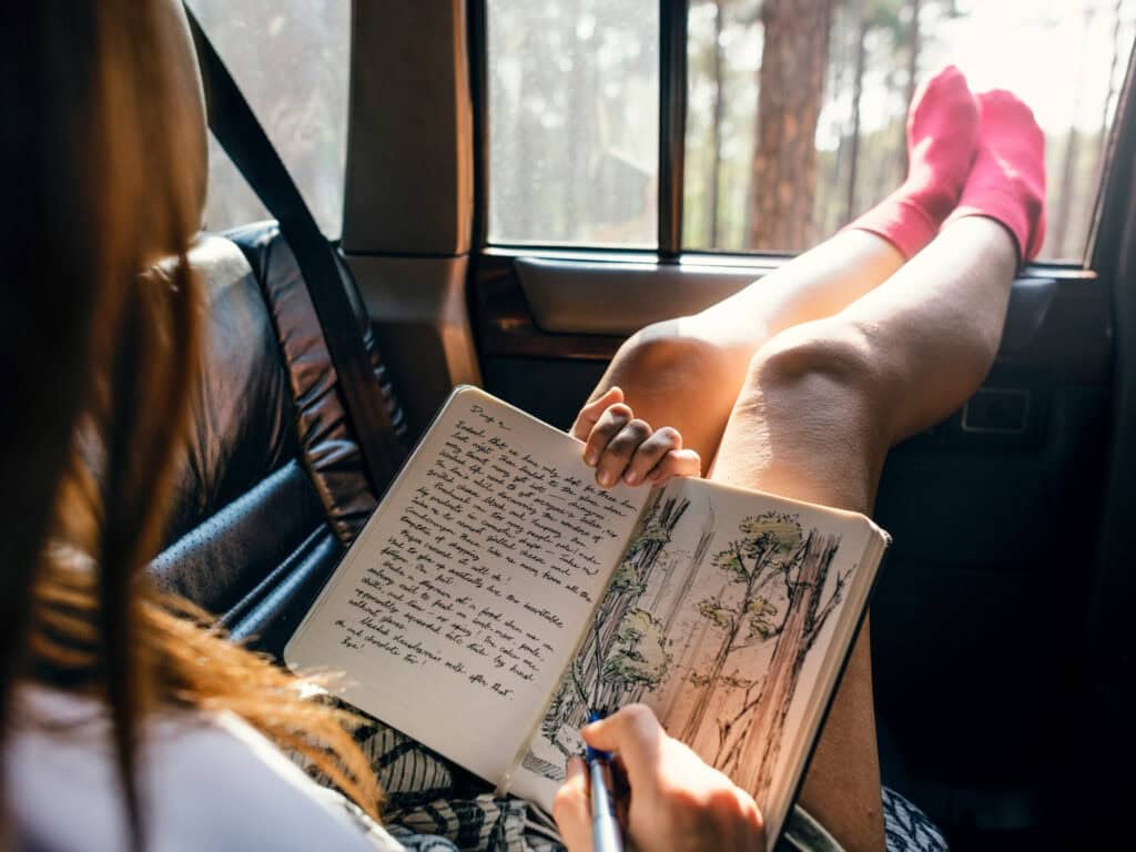 Eine Frau sitzt entspannt am Fenster mit Blick ins Grüne, die Füße hochgelegt. Sie schreibt in ein aufgeschlagenes Notizbuch mit handschriftlichem Text und einer gezeichneten Naturskizze. Natürliches Licht, ruhige Atmosphäre.