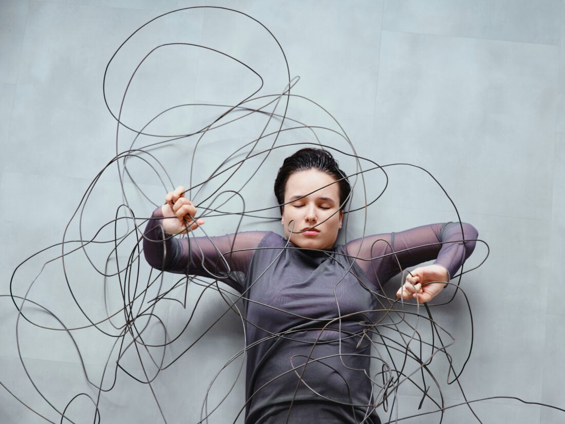 Frau liegt zwischen wirren, kabelartigen Linien, die sie umgeben und einengen – Symbol für wissenschaftlich klingende Erklärungen, die Orientierung versprechen, aber verwirren.
