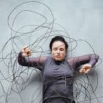 Frau liegt zwischen wirren, kabelartigen Linien, die sie umgeben und einengen – Symbol für wissenschaftlich klingende Erklärungen, die Orientierung versprechen, aber verwirren.