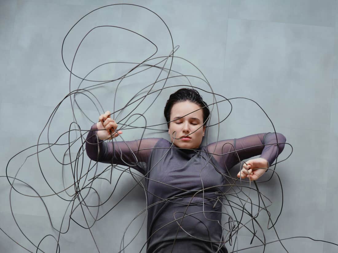 Frau liegt zwischen wirren, kabelartigen Linien, die sie umgeben und einengen – Symbol für wissenschaftlich klingende Erklärungen, die Orientierung versprechen, aber verwirren.