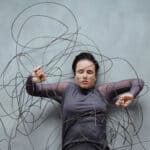 Frau liegt zwischen wirren, kabelartigen Linien, die sie umgeben und einengen – Symbol für wissenschaftlich klingende Erklärungen, die Orientierung versprechen, aber verwirren.