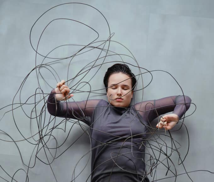 Frau liegt zwischen wirren, kabelartigen Linien, die sie umgeben und einengen – Symbol für wissenschaftlich klingende Erklärungen, die Orientierung versprechen, aber verwirren.