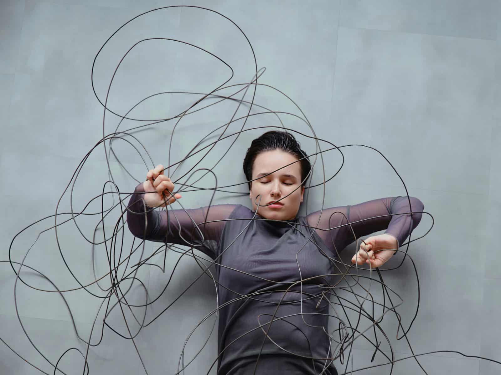 Frau liegt zwischen wirren, kabelartigen Linien, die sie umgeben und einengen – Symbol für wissenschaftlich klingende Erklärungen, die Orientierung versprechen, aber verwirren.