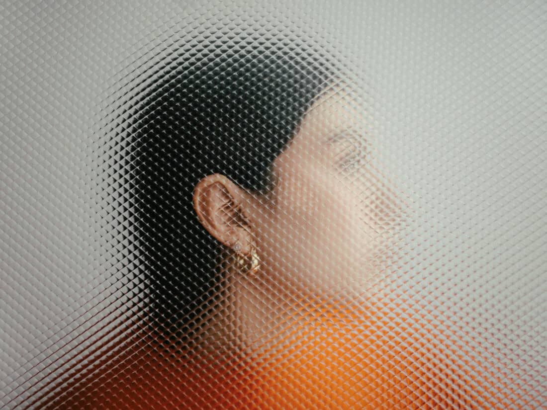 Frau im Profil hinter strukturiertem Glasfenster, Gesicht teilweise verzerrt, Blick nach außen. Symbolfoto für die Wirkung von Influencerinnen