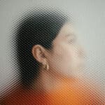 Frau im Profil hinter strukturiertem Glasfenster, Gesicht teilweise verzerrt, Blick nach außen. Symbolfoto für die Wirkung von Influencerinnen