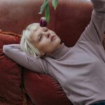 Frau liegt entspannt auf einer Couch und beschäftigt sich passiv mit einem Objekt; Darstellung einer von Influencerinnen geprägten, nach innen gerichteten Lebenshaltung ohne sichtbare Aktivität.