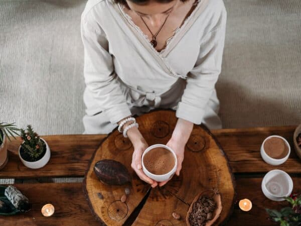 Person sitzt an einem Tisch und hält eine Tasse Kakao in den Händen, umgeben von Ritual-Objekten