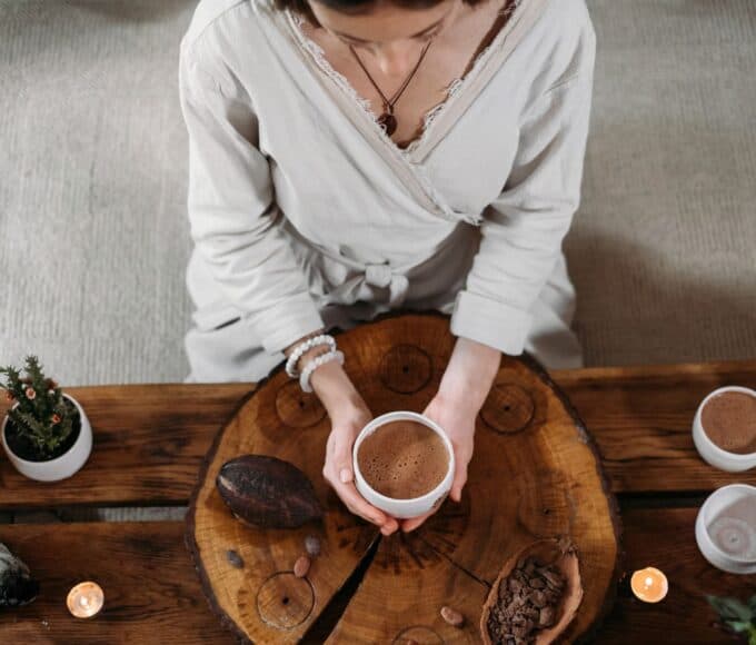 Person sitzt an einem Tisch und hält eine Tasse Kakao in den Händen, umgeben von Ritual-Objekten