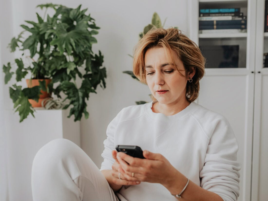 Frau sitzt in einem hellen Zimmer und schaut nachdenklich auf ihr Smartphone.