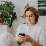 Frau sitzt in einem hellen Zimmer und schaut nachdenklich auf ihr Smartphone.