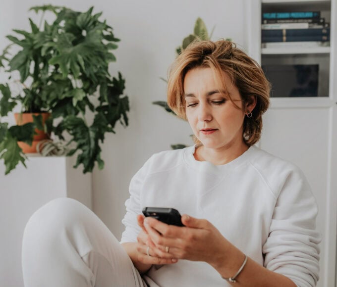 Frau sitzt in einem hellen Zimmer und schaut nachdenklich auf ihr Smartphone.