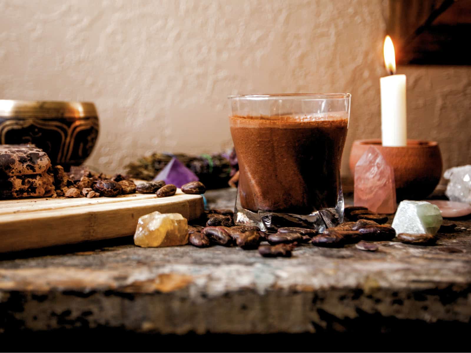 Ein Glas mit Kakao steht auf einem Tisch neben Kerze, Kristallen, Kakaobohnen und Kakaostücken in einem spirituell inszenierten Setting.