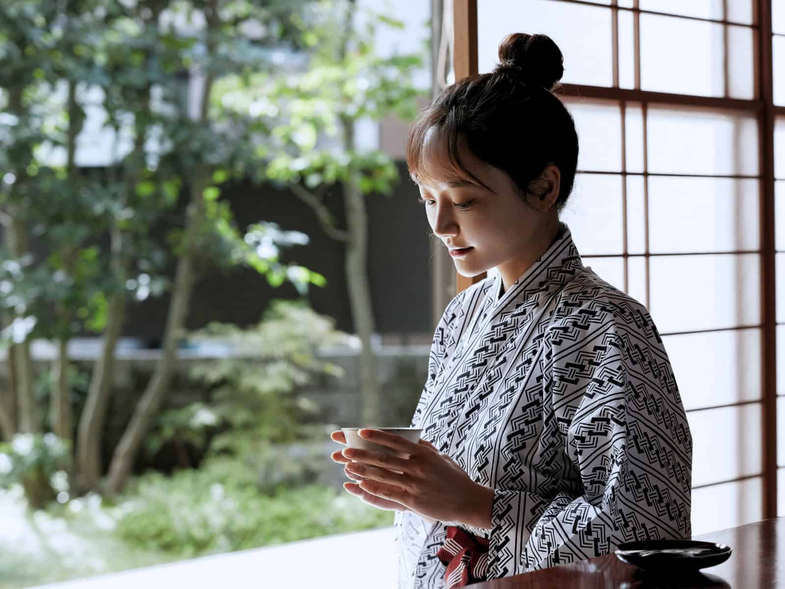 Eine Frau in einem traditionellen Kimono steht in einem japanischen Innenraum mit Schiebetüren und hält eine Tasse in den Händen.