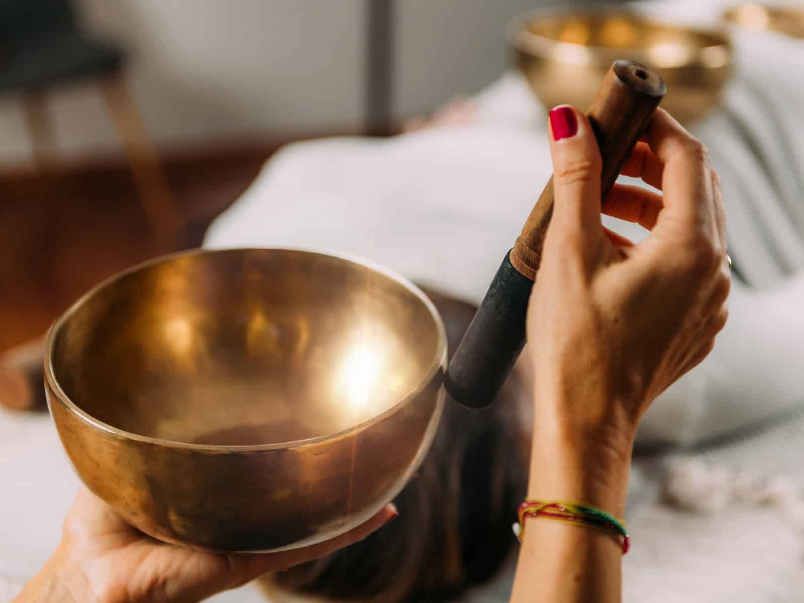 Eine Person hält eine Klangschale und einen Klöppel in einem ruhigen, wellnessartig inszenierten Setting.