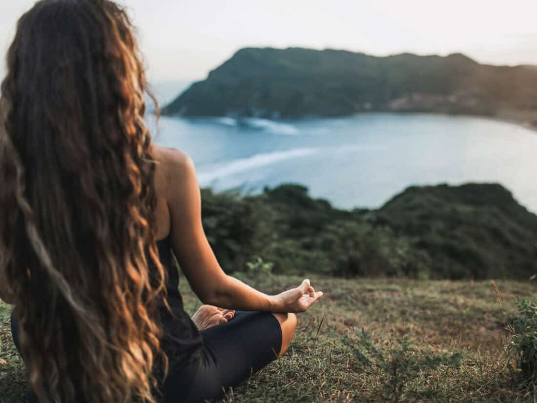 Frau sitzt im Schneidersitz auf einer Wiese und meditiert mit Blick auf eine Küstenlandschaft bei Sonnenuntergang.