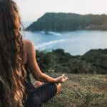 Frau sitzt im Schneidersitz auf einer Wiese und meditiert mit Blick auf eine Küstenlandschaft bei Sonnenuntergang.