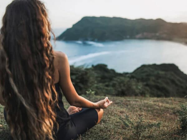 Frau sitzt im Schneidersitz auf einer Wiese und meditiert mit Blick auf eine Küstenlandschaft bei Sonnenuntergang.