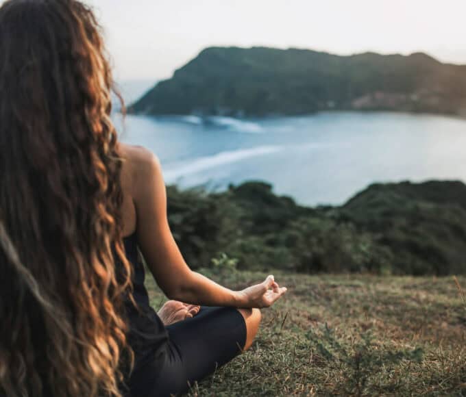 Frau sitzt im Schneidersitz auf einer Wiese und meditiert mit Blick auf eine Küstenlandschaft bei Sonnenuntergang.