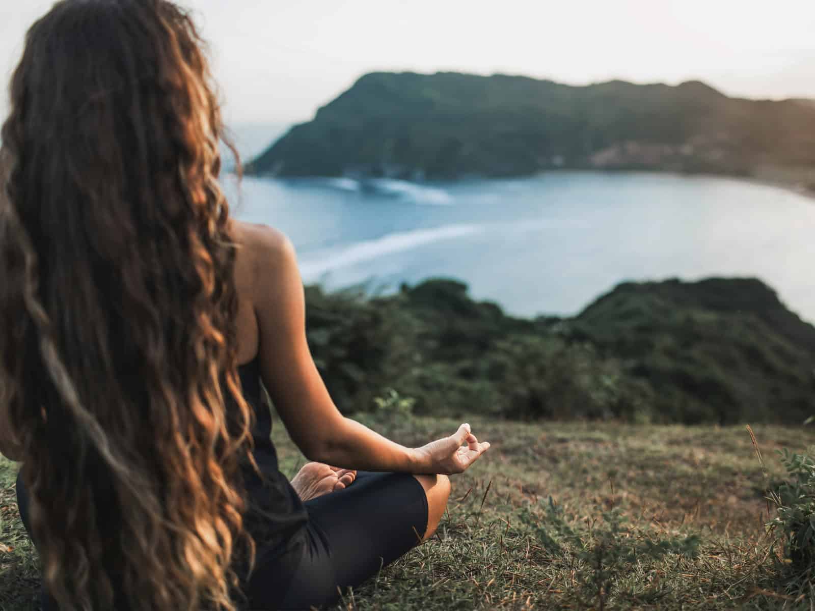 Frau sitzt im Schneidersitz auf einer Wiese und meditiert mit Blick auf eine Küstenlandschaft bei Sonnenuntergang.