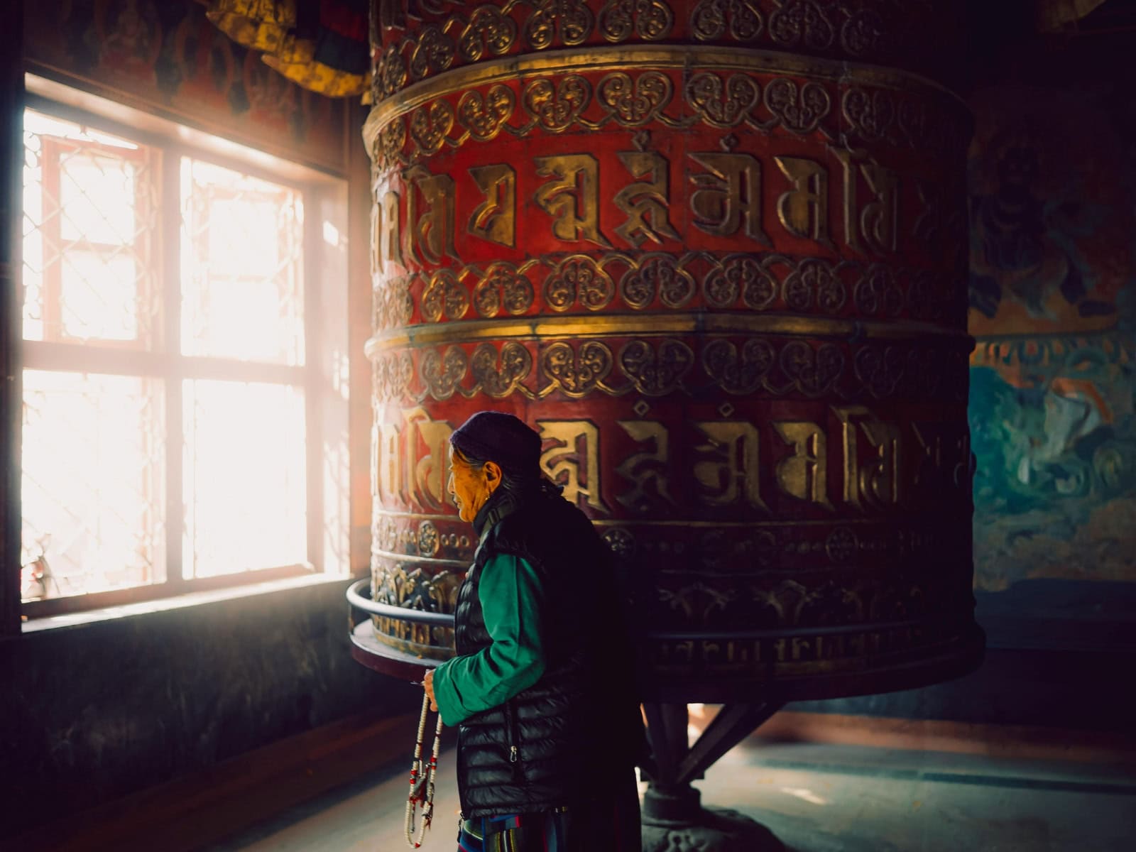 Eine Person steht in einem buddhistischen Kloster neben einer großen roten Gebetsmühle mit goldener Schrift und hält eine Gebetskette in der Hand.