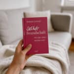 Eine Hand hält das pink-rote Buch „Selbstfreundschaft – Wie das Leben leichter wird“ von Wilhelm Schmid vor einem hellen Sofa mit cremefarbener Decke in einem ruhigen, weich unscharfen Wohnzimmer.