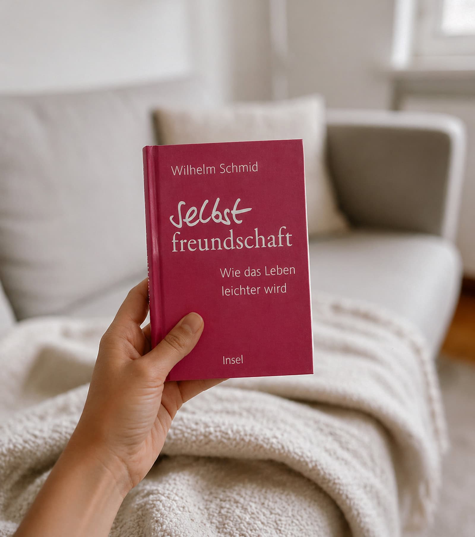 Eine Hand hält das pink-rote Buch „Selbstfreundschaft – Wie das Leben leichter wird“ von Wilhelm Schmid vor einem hellen Sofa mit cremefarbener Decke in einem ruhigen, weich unscharfen Wohnzimmer.