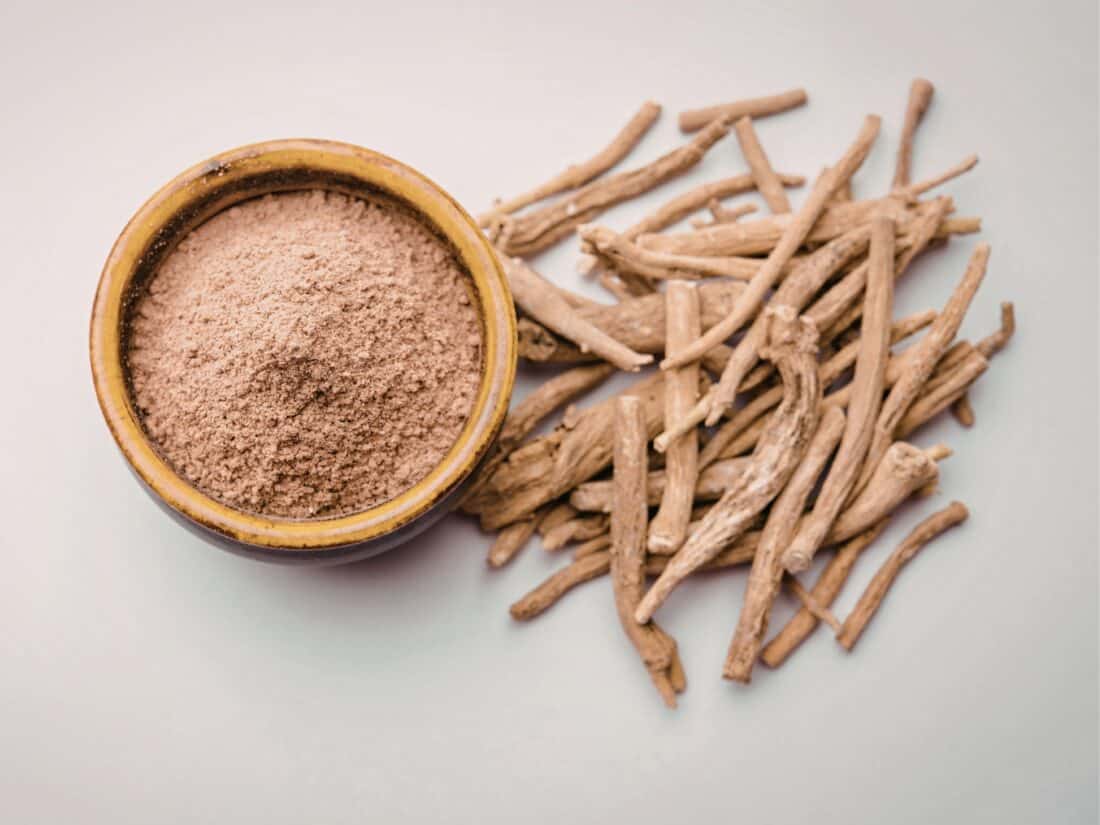 Ashwagandha-Wurzelstücke liegen neben einer Schale mit gemahlenem Ashwagandha-Pulver auf hellem Hintergrund.