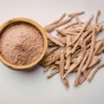 Ashwagandha-Wurzelstücke liegen neben einer Schale mit gemahlenem Ashwagandha-Pulver auf hellem Hintergrund.