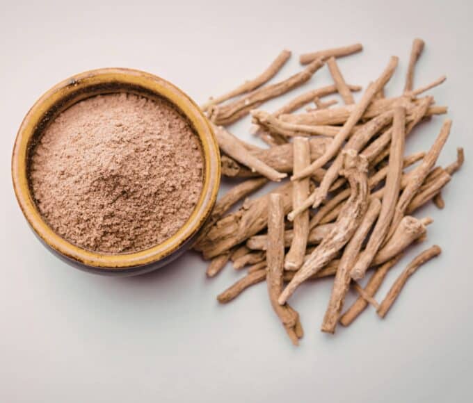 Ashwagandha-Wurzelstücke liegen neben einer Schale mit gemahlenem Ashwagandha-Pulver auf hellem Hintergrund.