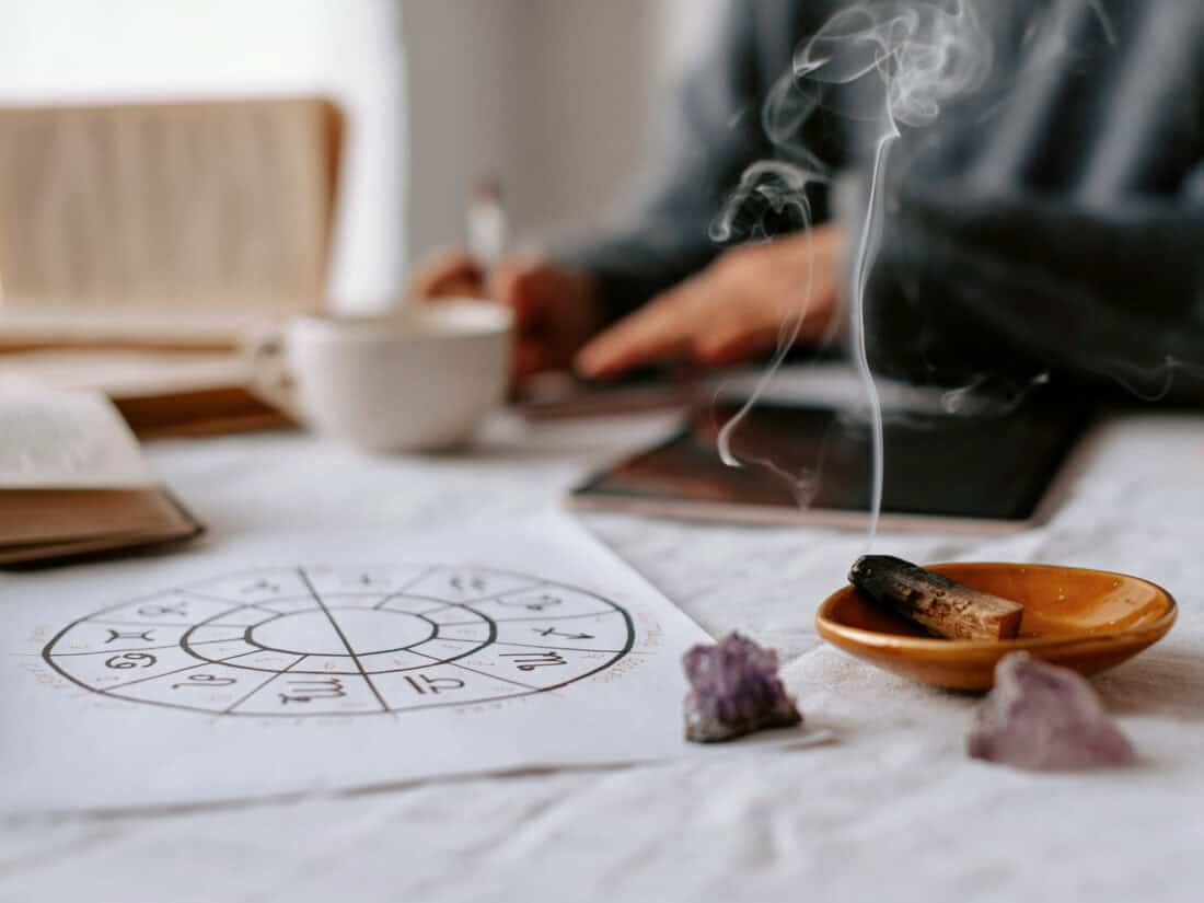 Astrologie-Zeichnung, Räucherholz und Amethyst-Steine auf einem hellen Tisch, im Hintergrund sitzt eine Person unscharf im meditativen Setting.