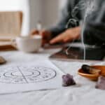 Astrologie-Zeichnung, Räucherholz und Amethyst-Steine auf einem hellen Tisch, im Hintergrund sitzt eine Person unscharf im meditativen Setting.