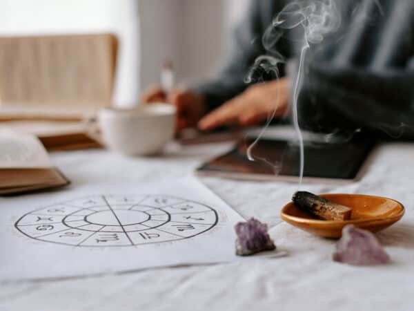 Astrologie-Zeichnung, Räucherholz und Amethyst-Steine auf einem hellen Tisch, im Hintergrund sitzt eine Person unscharf im meditativen Setting.