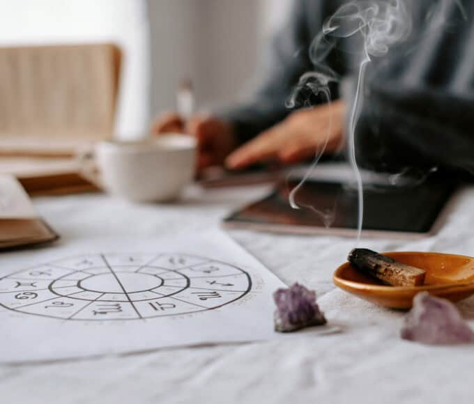 Astrologie-Zeichnung, Räucherholz und Amethyst-Steine auf einem hellen Tisch, im Hintergrund sitzt eine Person unscharf im meditativen Setting.