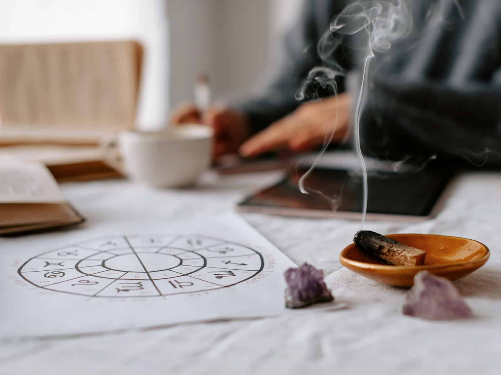 Astrologie-Zeichnung, Räucherholz und Amethyst-Steine auf einem hellen Tisch, im Hintergrund sitzt eine Person unscharf im meditativen Setting.