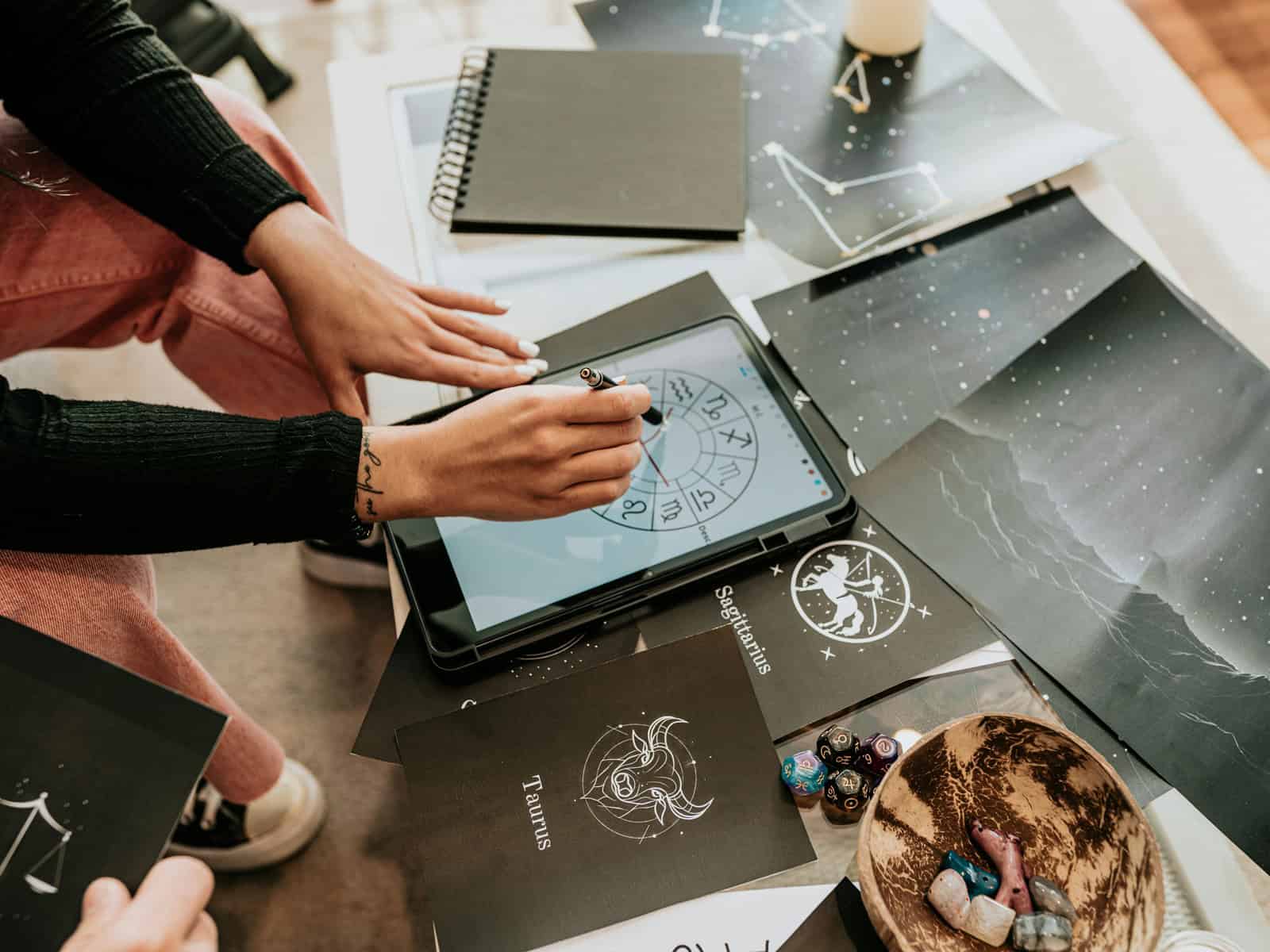 Modernes Esoterik-Setting mit Tablet, Sternzeichen-Grafiken, Kristallen, Karten und Ritualobjekten auf einem Tisch.