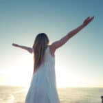 Frau in weißem Kleid steht mit ausgebreiteten Armen vor dem Meer und der aufgehenden Sonne als Symbolbild für moderne Spiritualität und Channeling