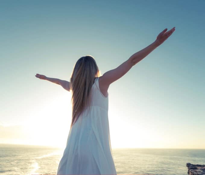 Frau in weißem Kleid steht mit ausgebreiteten Armen vor dem Meer und der aufgehenden Sonne als Symbolbild für moderne Spiritualität und Channeling