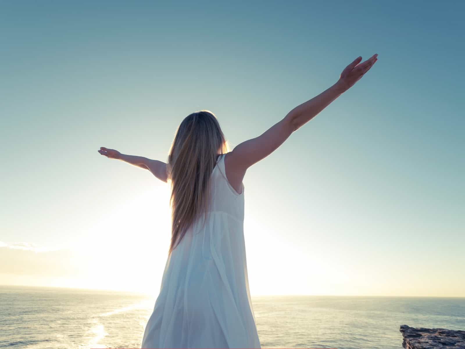 Frau in weißem Kleid steht mit ausgebreiteten Armen vor dem Meer und der aufgehenden Sonne als Symbolbild für moderne Spiritualität und Channeling